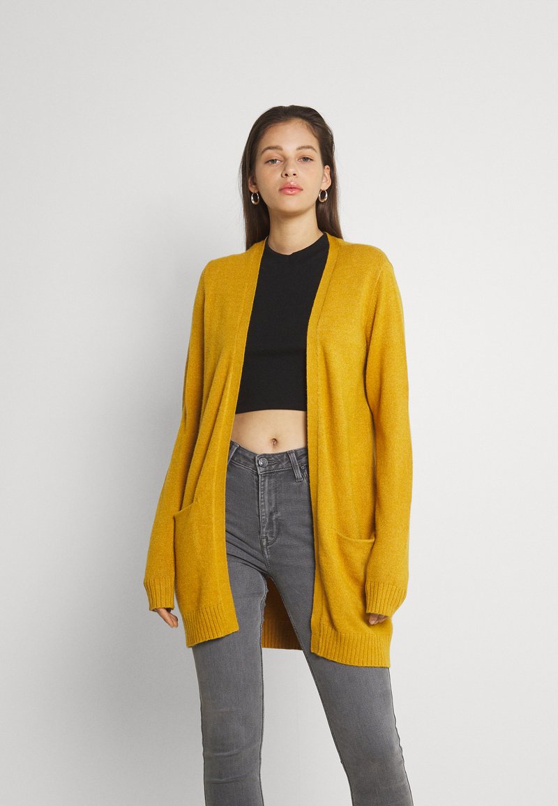 Cárdigan amarillo de punto con abertura frontal, mangas largas, puños acanalados y dos bolsillos frontales. Llevado sobre un top corto negro y unos jeans grises.
