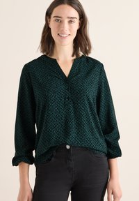 Dunkelgrüne Bluse mit geometrischem Muster in Petrol, V-Ausschnitt und langen Ärmeln mit hochgerollten Bündchen. Kombiniert mit schwarzen Jeans.