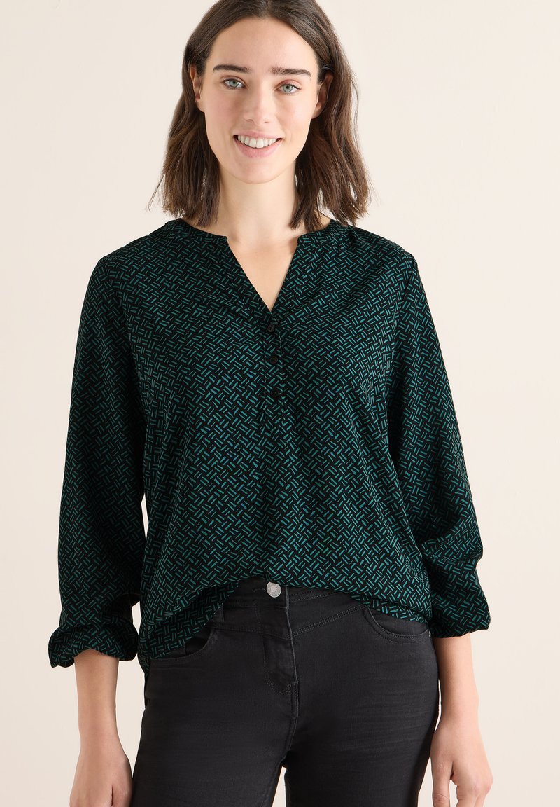Dunkelgrüne Bluse mit geometrischem Muster in Petrol, V-Ausschnitt und langen Ärmeln mit hochgerollten Bündchen. Kombiniert mit schwarzen Jeans.