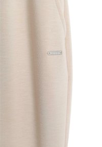 Lys beige sweatshirtstoff med en glatt tekstur og sidelomme. Har et sølvfarget metalllogo som detalj. Enkel, minimalistisk design.