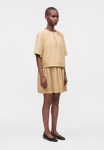 Femme debout portant un haut beige à manches courtes et une jupe assortie avec un ourlet à motif, associée à des chaussures plates noires, sur un fond uni.