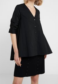 Svart knapplöst skjorta med V-ringning, lös passform och uppvikta ärmar, tillsammans med en svart kjol med vertikala utsmyckningar.