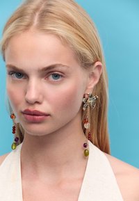 Goldfarbene Ohrringe mit floralem Design, die bunten Perlen in Lila, Rot, Orange und Grün aufweisen und an zarten Ketten hängen.