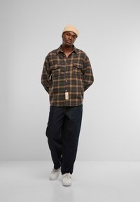 Geruite flanellen overhemd in donker groen, bruin en gele tinten. Twee borstzakken en een label aan de onderkant. Draagt met donkere jeans en witte schoenen.