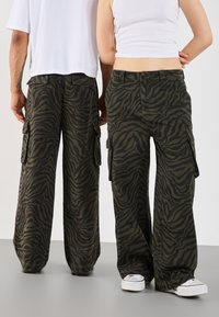 Cargo pants in olive green with a black zebra stripe pattern, featuring side pockets and a relaxed fit. Worn with white sneakers.