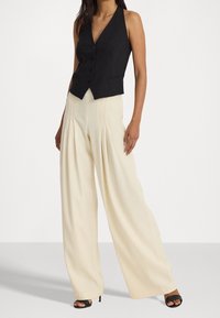 Nainen, jolla on päällään musta napillinen liivi, jossa on poolokaulus, sekä korkeavyötäröiset, leveälahkeiset vaaleat housut, joissa on laskokset. Mustat avokkaat.