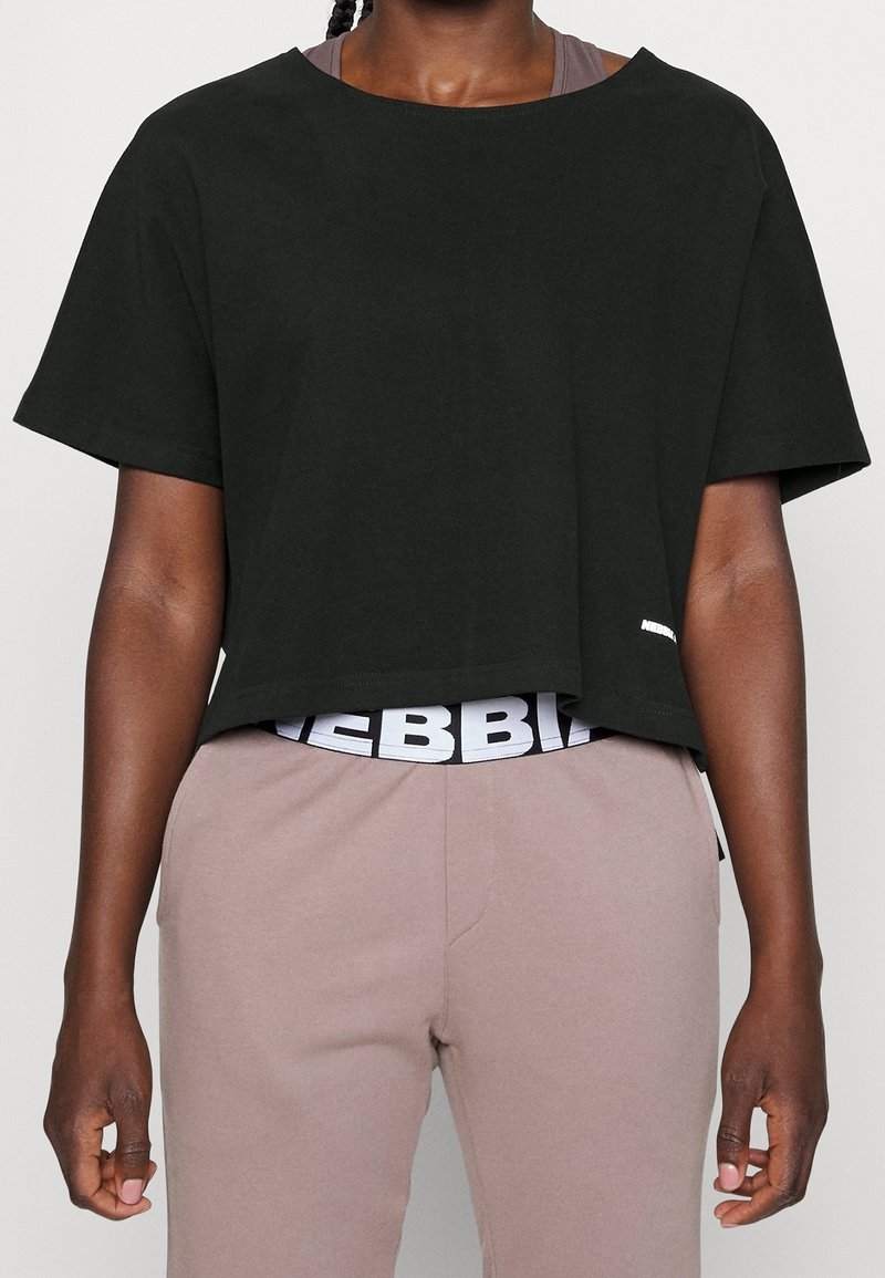 Personne portant un crop top noir à manches courtes sur un pantalon taupe avec une ceinture noire et blanche visible en dessous.