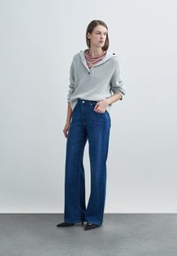 Ανοιχτό γκρι πλεκτό πουλόβερ με ψηλό λαιμό και διακοσμητικά κουμπιά, συνδυασμένο με μπλε τζιν παντελόνια με φαρδύ πόδι και μαύρα μυτερά παπούτσια.