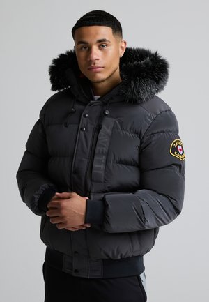 Ung mand iført en sort dunjakke med pelsforet hætte og et Canada-patch på ærmet, stående mod en ensfarvet baggrund.