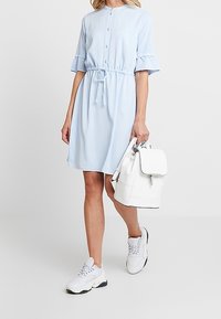 Jasnoniebieska bawełniana sukienka z guzikiem z przodu, krótkimi falbaniastymi rękawami i pasem ze sznurkiem, w zestawieniu z białym plecakiem i masywnymi białymi sneakersami.