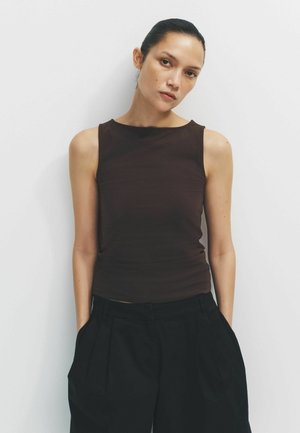 Naine tumedate tagasitõmmatud juustega, kandes varrukateta tumedat pruunikat toppi ja musti pükse, seisab valge seina vastas käed taskutes.