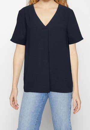 Vrouw met een effen donkerblauw T-shirt met korte mouwen en V-hals, en lichtblauwe spijkerbroek, staand tegen een effen lichte achtergrond.