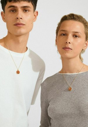 Due adulti in posizione frontale, che indossano collane d'oro con pendenti rotondi arancioni, uno con una camicia bianca e l'altro con un top grigio a coste.