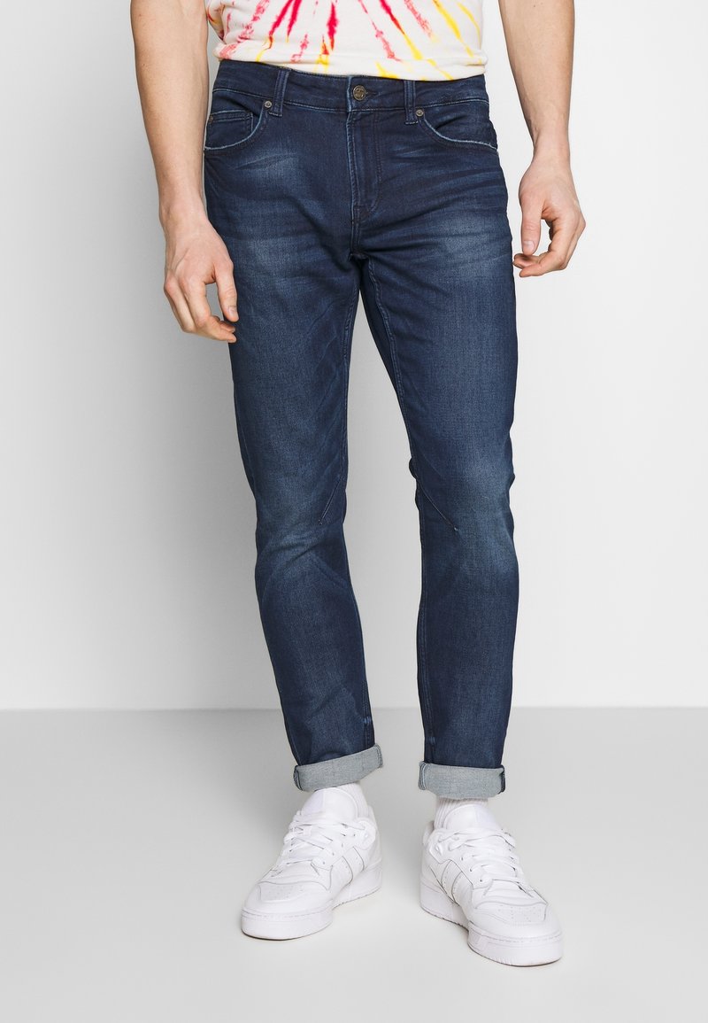 Dark blue denim jeans with a slim fit, visible stitching, rolled cuffs, and front/back pockets. Worn with white sneakers and a tie-dye shirt.