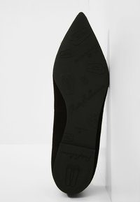 Chaussure plate noire à bout pointu avec semelle texturée présentant des motifs de chaussons de ballet et un logo sur fond blanc.