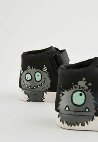 Scarpe nere alte con design di mostri dei cartoni animati, con pelliccia grigia e occhi blu. Include una fascia in velcro per una vestibilità sicura. Suola bianca.