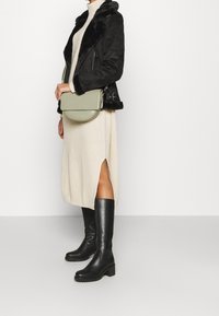 Giacca nera in suede con finiture in pelliccia sintetica, abito in maglia crema con spacco laterale, borsa a tracolla verde, stivali neri in pelle alti con tacco robusto.