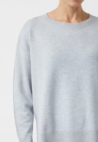 Oberkörper, der einen hellgrauen Strickpullover mit geripptem Rundhalsausschnitt, Bündchen und Saum trägt, vor einem schlichten weißen Hintergrund.