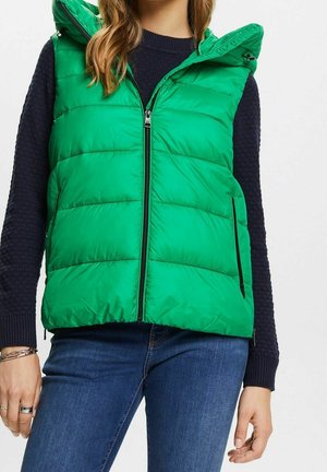 Femme portant un gilet matelassé sans manches vert vif sur un pull tricoté foncé et un jean bleu, avec des bracelets et des bagues à la main gauche.