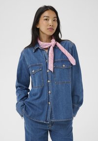 Camicia di denim blu medio, con due tasche sul petto, bottoni argentati e una vestibilità morbida. Abbinata a una sciarpa rosa con motivo attorno al collo.