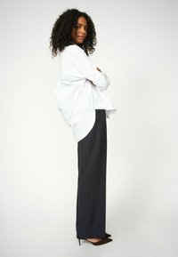 Weißes, übergroßes Hemd mit abgerundetem Saum, kombiniert mit maßgeschneiderten, dunklen Hosen und braunen, spitz zulaufenden Heels. Lockige Haare offen gestylt.