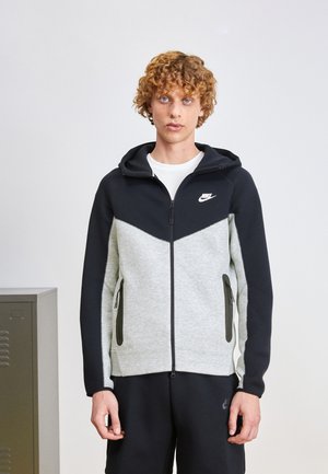 Felpa Nike con zip in grigio e nero, caratterizzata da un motivo a chevron, tasche laterali con zip e cappuccio regolabile con cordino. Materiale misto cotone.