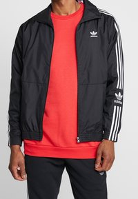 Svart vindjacka med vita ränder på ärmarna, med dragkedja och elastiska manschetter. Buren över en röd sweatshirt och svarta byxor.