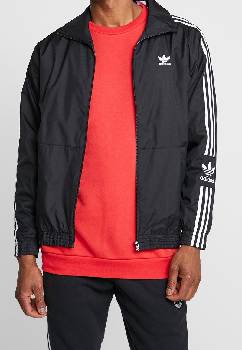 Svart vindjacka med vita ränder på ärmarna, med dragkedja och elastiska manschetter. Buren över en röd sweatshirt och svarta byxor.