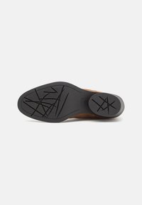Zapato de ante marrón con una suela de goma negra estructurada que presenta un patrón de agarre texturizado. Diseño de punta redonda y tacón plano.