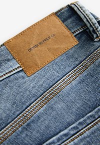 Jeanid, millel tõukereha nahakott, millel on kiri "DENIM SUPPLY Co." Sinine kude sisaldab kontrastset tan värvi õmblust ja peent tekstuuri.