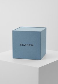 Mėlyna kvadratinė prezentacinė dėžutė, pagaminta iš tekstūruoto popieriaus, su juodu "SKAGEN" prekės ženklu vienoje pusėje ir plokščiu dangčiu.