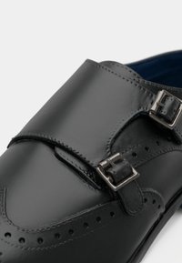 Černé kožené boty s lesklým povrchem, vybavené dvojitými stříbrnými přezkami, dekorativními perforacemi podél okrajů a texturovanou stélkou.