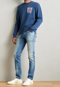 Granatowa bluza z różowym nadrukiem, do tego jasnoniebieskie obcisłe dżinsy i białe trampki z beżowymi akcentami, stojące na drewnianej podłodze.