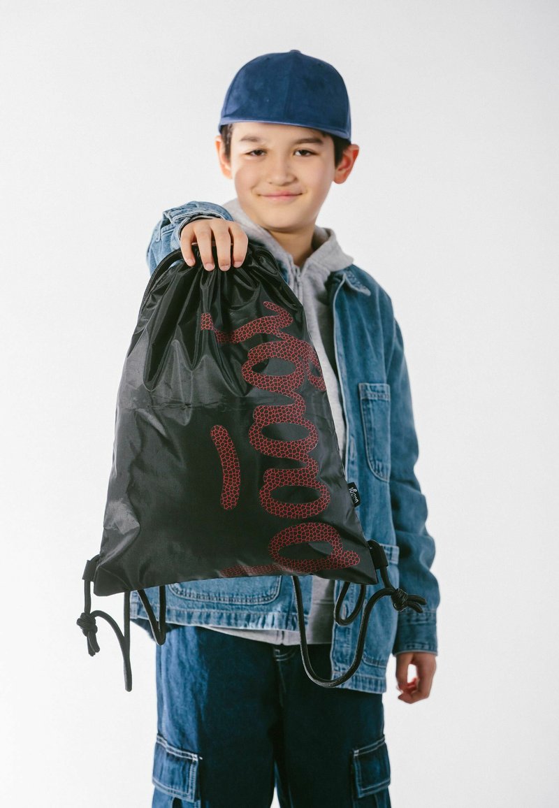 Schwarze Zugtasche mit rotem Grafikdesign, aus leichtem Material, gehalten von einem Kind, das eine Jeansjacke und einen grauen Kapuzenpullover trägt.