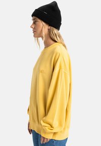 Gele oversized sweatshirt met ribbelmanchetten, casual fit, met een geborduurd logo. Gecombineerd met blauwe spijkerbroek en een zwarte beanie.