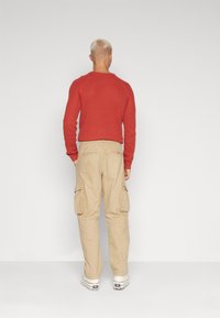 Rød ribstrikket sweater, cargo-bukser i tan med sidelommer og hvide sneakers med sorte accenter. Ensfarvet baggrund, set bagfra.