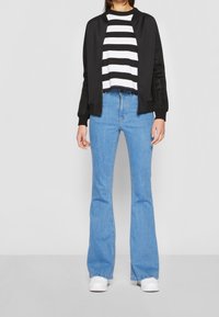 Černá bomber bunda s síťovými akcenty, bílo-černý pruhovaný crop top, světle modré zvonové džíny a bílé tenisky.