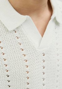Vit, stickad polo med en texturerad design har en V-hals och små hål som skapar ett mönster, vilket framhäver ett mjukt och lätt material.