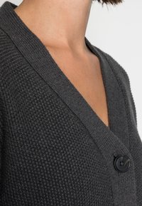 Tmavě šedý texturovaný kardigan s výstřihem do V, zdobený tkaným vzorem a jedním černým knoflíkem. Měkká látka a minimalistické designové detaily.