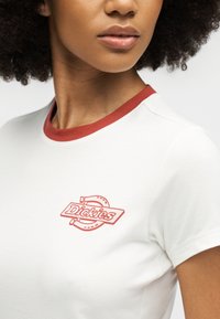 Γυναίκα που φοράει λευκό μπλουζάκι με κόκκινο γιακά και μικρό κόκκινο λογότυπο "Dickies" στο στήθος, με σγουρά μαλλιά και ουδέτερο χρώμα χειλιών.