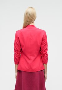 Blazer rosso su misura con maniche arricciate, caratterizzato da un design strutturato e tessuto liscio. Indossato sopra una gonna bordeaux abbinata.