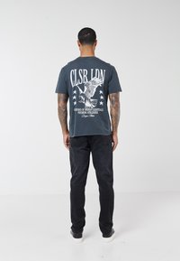 Muškarac s tetovažama stoji okrenut leđima, nosi tamnosivu grafičku majicu s pticom i tekstom, crne traperice i crno-bijele tenisice.