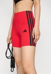 Crvene atletske kratke hlače od rastezljivog materijala, s crnim bočnim trakama i Adidas logotipom na lijevoj nozi. Lagane i prilagodljive.