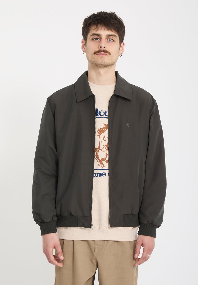 Jeune homme avec moustache portant une veste noire zippée sur un t-shirt graphique beige et un pantalon kaki, debout devant un fond blanc uni.