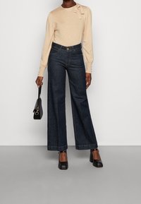 Kvinna i beige långärmad topp med rosett, högmidjade vida mörkblå jeans, svart handväska och svarta klackskor med blockklack.