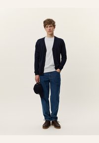 Cardigan blu navy con chiusura a bottoni, t-shirt grigia chiara, jeans in denim blu larghi e scarpe marroni. Tiene un cappellino nero. Look semplice e casual.
