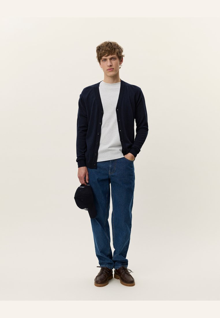 Cardigan blu navy con chiusura a bottoni, t-shirt grigia chiara, jeans in denim blu larghi e scarpe marroni. Tiene un cappellino nero. Look semplice e casual.
