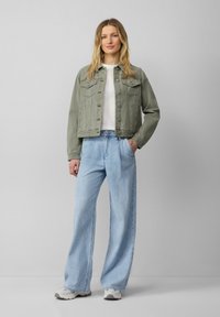 Przycięta zielona kurtka dżinsowa na białej koszulce, połączona z jasnoniebieskimi spodniami typu wide-leg oraz szarymi sneakersami. Luźny, codzienny zestaw.