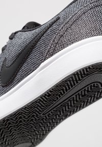 Grå mesh-sko med svarta detaljer, vit mellansula och mönstrad gummiyttersula med geometriskt mönster.
