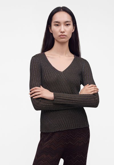 Femme aux longs cheveux foncés portant un pull noir côtelé à col en V et un pantalon sombre à motif zigzag, debout les bras croisés devant un fond blanc.